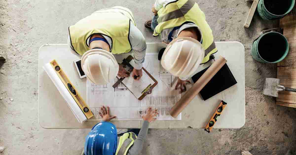 A trio of people in hard hats leaning over a table with a floor plan, a clipboard, and drafting tools.