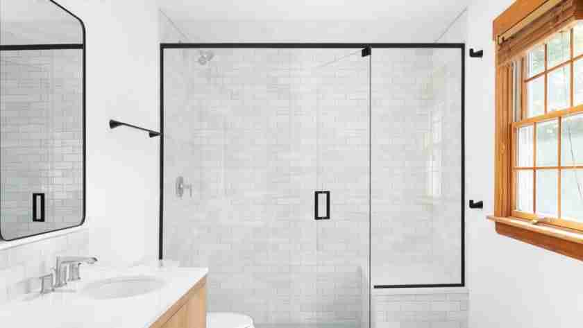 A luxury walk-in shower with white tile, glass doors, and black trim in a clean, white bathroom.