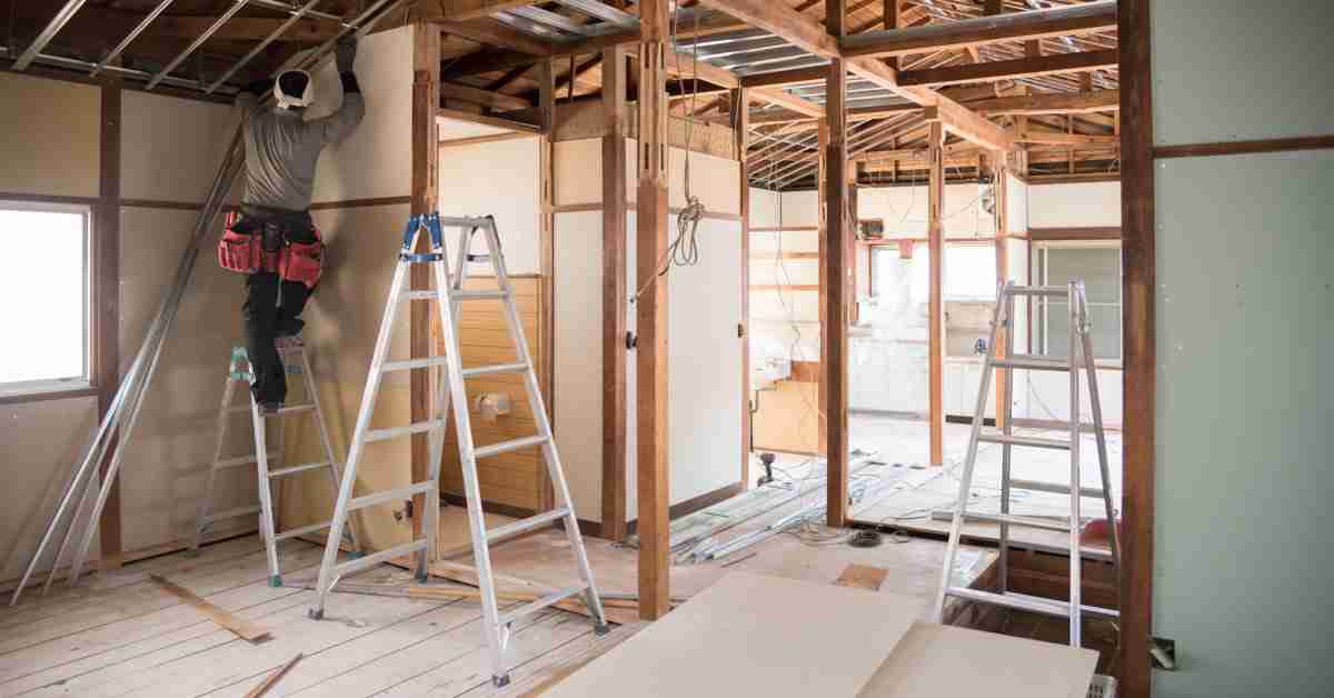 A room undergoing construction, with unfinished floors and exposed beams. A construction worker stands on a ladder in the corner.