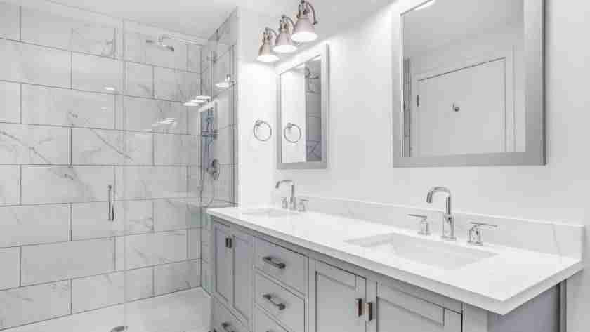 A white, minimalist bathroom with gray cabinets, white tile, white countertops, and glass shower walls.