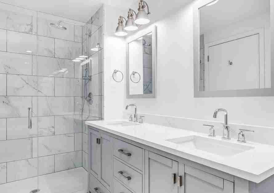A white, minimalist bathroom with gray cabinets, white tile, white countertops, and glass shower walls.