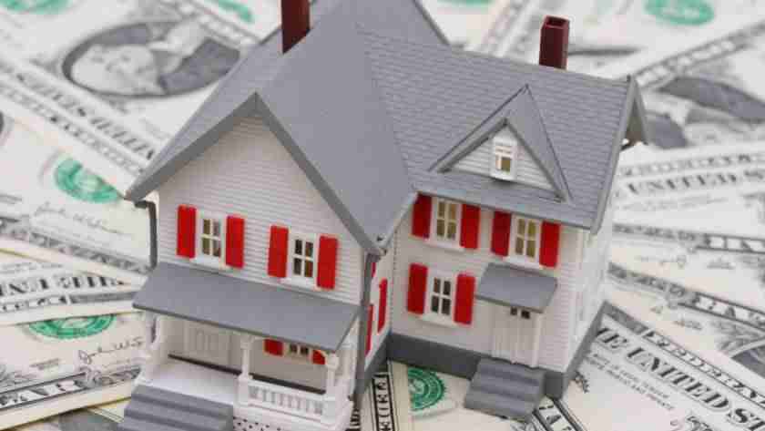 A small model of a two-story house sitting on a stack of one dollar bills that fan out in a circular shape beneath it.
