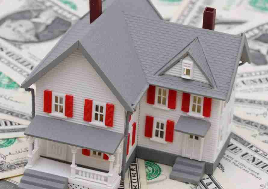 A small model of a two-story house sitting on a stack of one dollar bills that fan out in a circular shape beneath it.